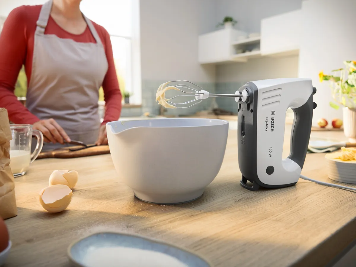 Eine Person mit Schürze bereitet sich darauf vor, einen elektrischen Handmixer zu benutzen; daneben stehen eine Rührschüssel und aufgeschlagene Eier auf einer Küchenarbeitsplatte aus Holz.