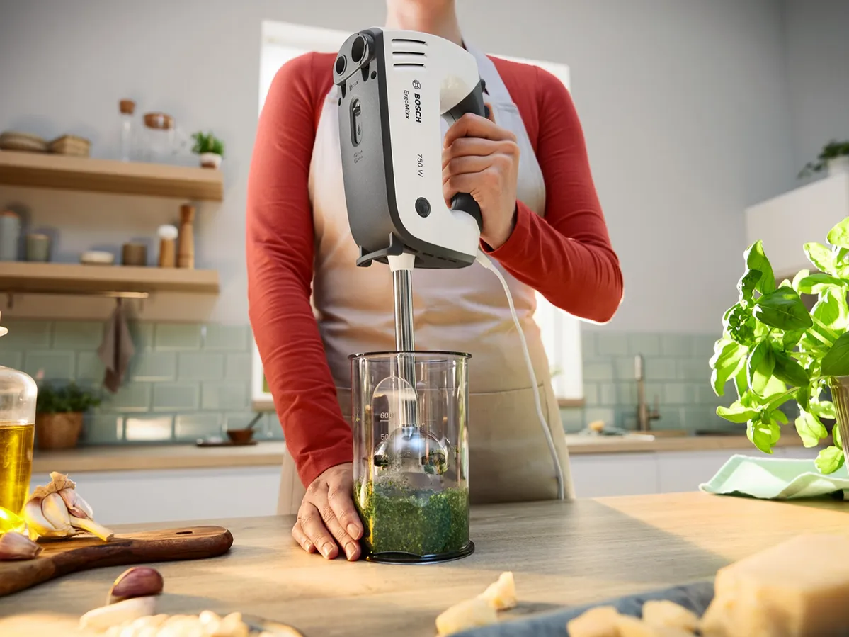 Eine Person verwendet einen Stabmixer in einem Glasbehälter, um grüne Kräuter in einer hellen Küche zu pürieren.