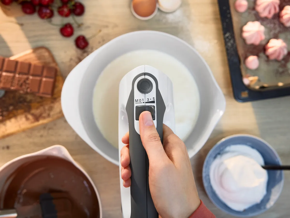 Hand hält einen weißen Stabmixer über einer Schüssel mit weißer Flüssigkeit auf einem Holztisch mit Schokolade, Kirschen und weiteren Schüsseln im Hintergrund