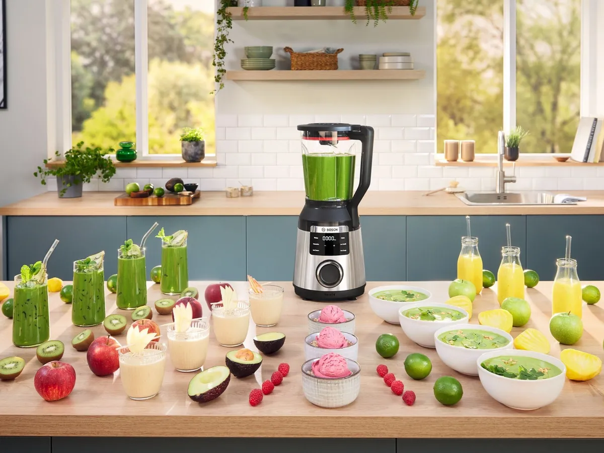 Küchenarbeitsplatte mit einem Bosch-Standmixer, umgeben von grünen Smoothies, gelben Säften, Obst, Suppenschalen und rosafarbenen Eiskugeln.