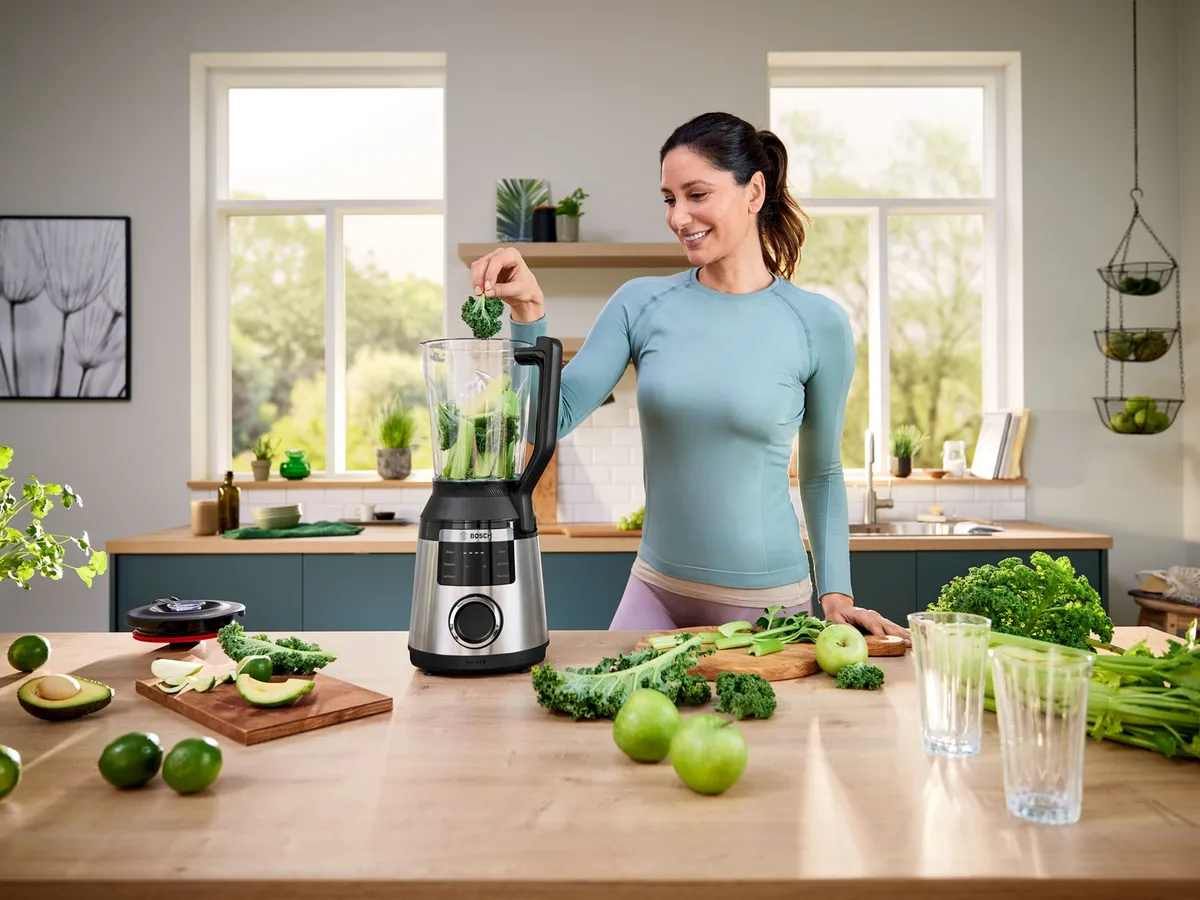 Eine Frau gibt Grünkohl in einen Mixer; sie befindet sich in einer hellen, modernen Küche, auf deren Arbeitsplatte grünes Gemüse und Obst liegen.