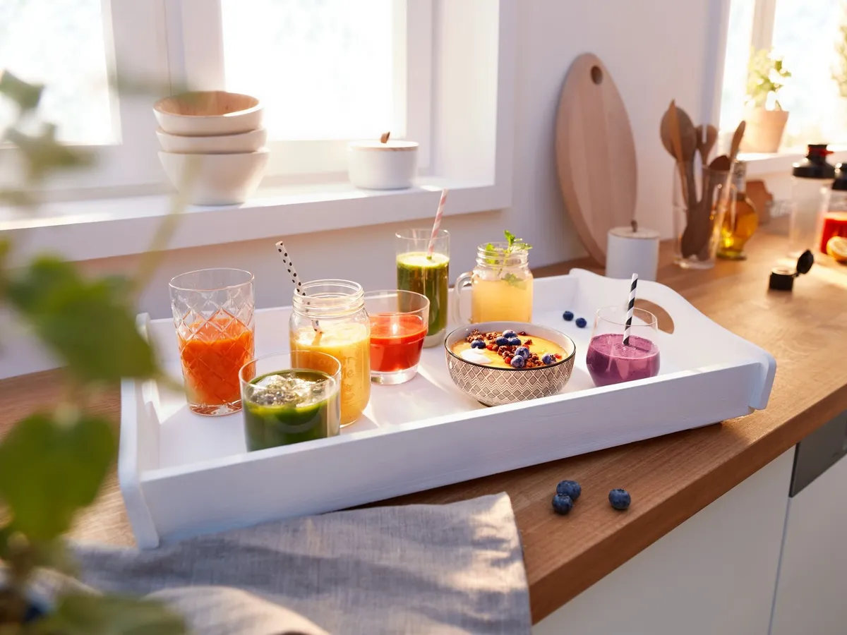 Ein weißes Tablett mit bunten Smoothies und einer Smoothie-Bowl mit Blaubeeren auf einer hölzernen Küchenarbeitsplatte am Fenster.