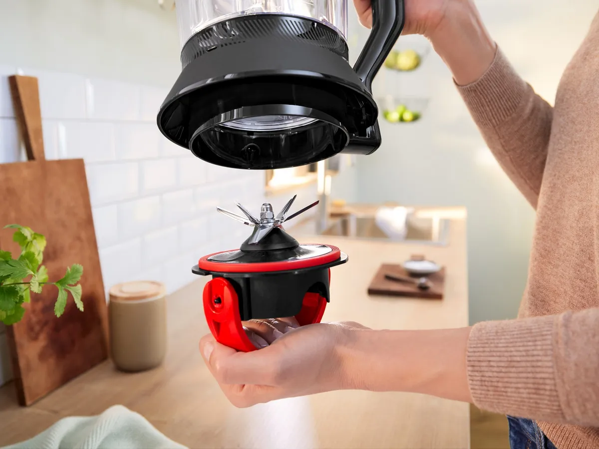 Person hält ein scharfes Mixermesser in der Hand, das in einer hellen Küchenumgebung aus einem Mixerbehälter entnommen wird.