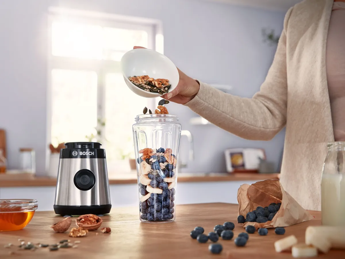 Here's an example:  Hand pouring nuts into a Bosch personal blender cup filled with blueberries and bananas on a kitchen counter.