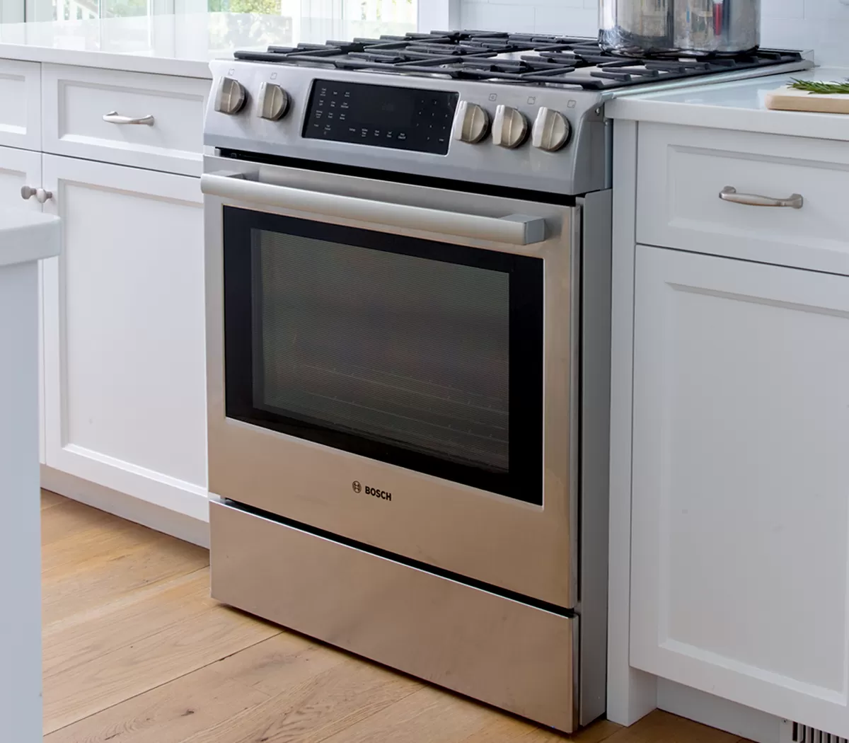 A sleek stainless steel Bosch gas range with a glass oven door, set against white cabinets and light wooden flooring.