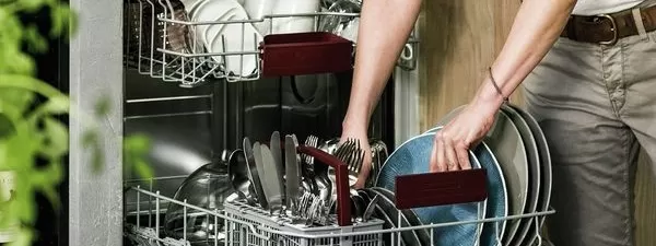 Eine Person entnimmt sauberes Geschirr aus einem Geschirrspüler und ordnet Teller und Utensilien mit zwei roten Geschirrkörben in das Gerät ein.