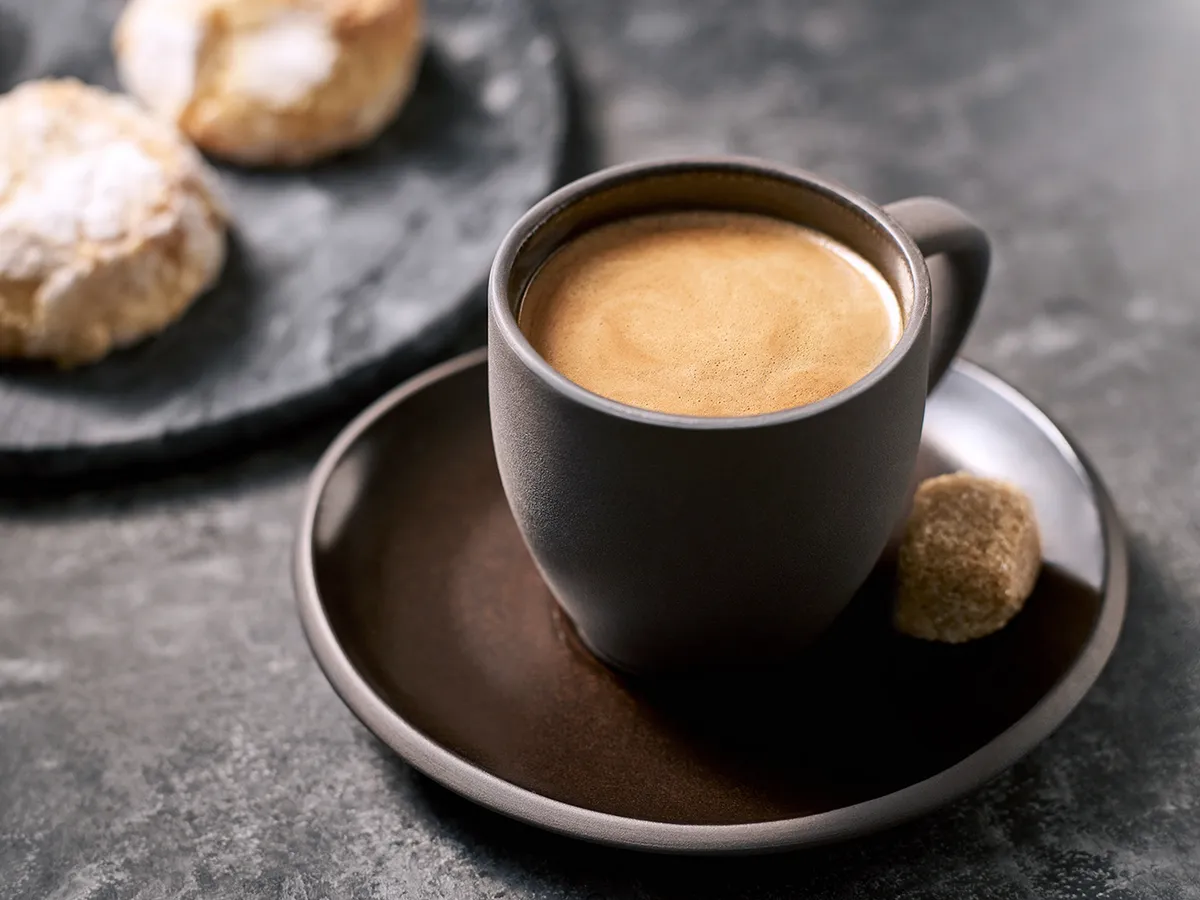 A steaming cup of coffee on a dark saucer, accompanied by a sugar cube, with fluffy pastries blurred in the background.