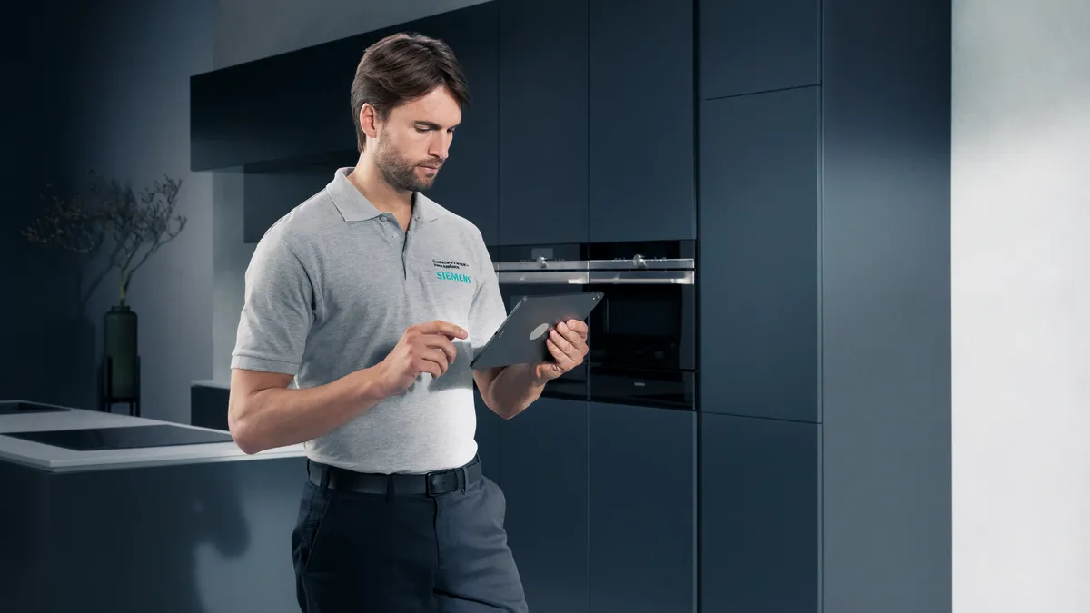 Техник в сив поло на Siemens стои в съвременна кухня, държи таблет и преглежда уредите в съответствие с елегантните шкафове.