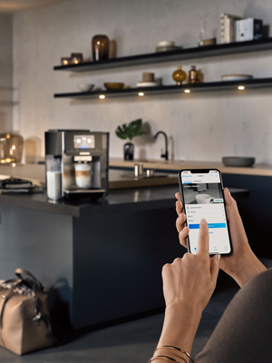 Kaffeevollautmat im Bildhintergrund mit Wohnlandschaft. Ein Hand mit Smart Phone im Vordergrund