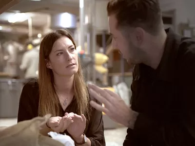 A man animatedly talks to a woman in a professional kitchen setting.
