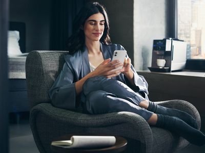 Link öffnet Infoseite über Home Connect für Siemens Hausgeräte; eine Frau bedient einen Siemens Kaffeevollautomaten via Smartphone