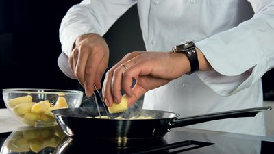 Uno chef aggiunge ingredienti a una padella sfrigolante mentre prepara un piatto, con una ciotola di patate tagliate accanto.