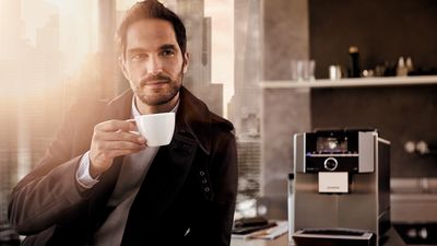 Siemens Kaffeewelt - Ein Mann trinkt Kaffee in der Küche. Ein Siemens EQ.9 Kaffeevollautomat steht auf dem Tisch.