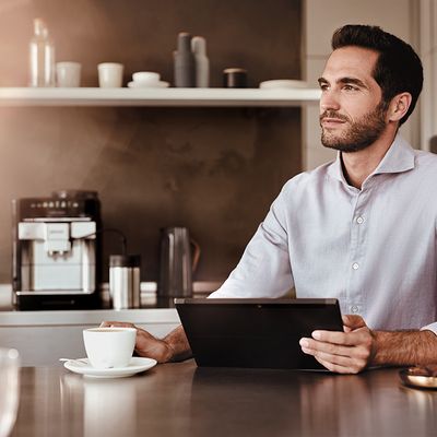 Ein Mann sitzt mit einer Tasse Kaffee in der Küche, ein EQ.6 plus Kaffeevollautomat im Hintergrund