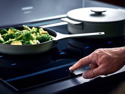 Siemens: eine Hand schaltet das Kochfeld ein, auf dem sich ein Topf mit Gemüse befindet