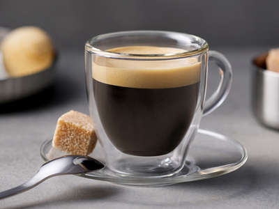 Un café expresso dans une tasse en verre avec une cuillère et un morceau de sucre à côté.