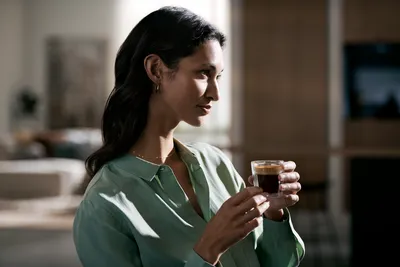 A woman holds a glass of dark espresso with thick cream in her hands.