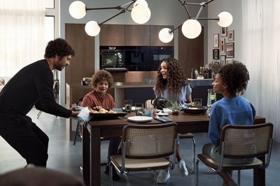 משפחה של אבא, אמא, בת ובן יושבים בשולחן עם אוכל עליו, האבא מגיש תבנית עם מאפים שהוציא מתנור חכם של סימנס