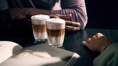 Arms and hands of two people on a table, sitting across from each other. Between them two full cappuccino glasses.