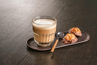Une tasse transparente contenant un flat white, pos&eacute;e sur un petit plateau individuel, accompagn&eacute; de  mignardises.