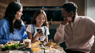 A man and woman sit at a table with a girl between them. She is smiling whilst showing them something on their phone.