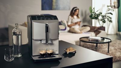 A sleek coffee machine sits on a dark table, two cups of espresso ready, with a water bottle and snacks beside it in a cozy living space.