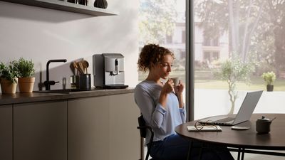 Kvinde, der sidder ved et bord i et moderne køkken og drikker en kop kaffe, mens hun kigger på sin laptop