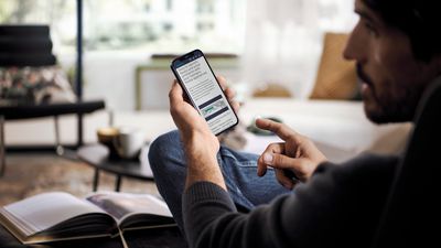 Un hombre sentado consulta información sobre su electrodoméstico en su teléfono móvil, señalando la pantalla con el dedo. Está en el salón y muestra un ambiente relajado en casa.