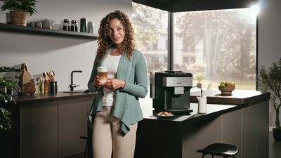 Smiling woman with a latte macchiato in her hands standing in front of a kitchen island; the EQ500 is placed next to her.
