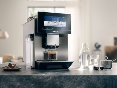Ein EQ900 Kaffeevollautomat steht auf einer Granitarbeitsplatte, daneben eine Wasserkaraffe mit einem Glas, Gebäck auf einem kleinen Teller und ein Zuckerschälchen.