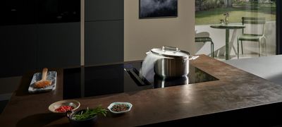 A black Siemens induction cooktop with an integrated downdraft extractor actively pulling steam from a pot, set in a modern kitchen with a view to an outdoor patio.