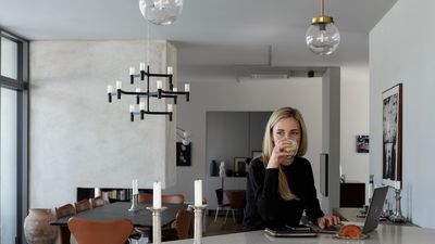 A woman sitting at a table in a modern home, working on a laptop with a cup of coffee beside her.