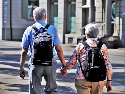 Para seniorów z plecakami spaceruje ulicą miasta, trzymając się za ręce.