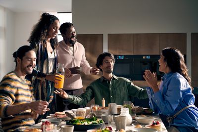 A diverse group of five friends enjoys a lively brunch or dinner around a table laden with food, laughing and interacting in a modern kitchen setting.