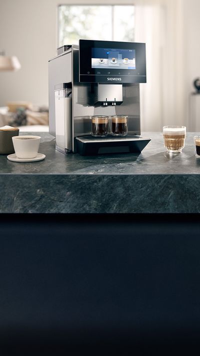 Ein Siemens EQ900 Kaffeevollautomat mit beanIdent System stimmt alle Geräteeinstelllungen auf Dallmayr und EQ Kaffeebohnen ab und bereitet so perfekte Kaffeespezialitäten zu. Er steht hier auf der Arbeitsplatte einer Küche. Um ihn herum stehen verschiedene Kaffeespezialitäten.