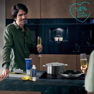 Ein Mann steht in der Küche an der Arbeitsfläche mit einem Glas Sekt in der Hand. Auf der Arbeitsfläche stehen Siemens Reinigungsmittel fü Hausgeräte. Es ist ein Störer zu sehen mit der info auf -14% Rabatt im Siemens Shop.