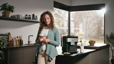 Femme souriante, un latte macchiato à la main, se tient debout devant un îlot de cuisine ; la machine EQ.500 est placé à côté d'elle.