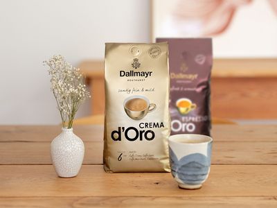 Es stehen eine goldene Packung Dallmayr Crema Kaffeebohnen und eine braune Packung Dallmayr Espresso d'Oro auf einem Tisch. Davor steht ein Kaffeebecher in Blau-Weiß. Daneben steht eine kleine Vase mit einem Sträußchen.