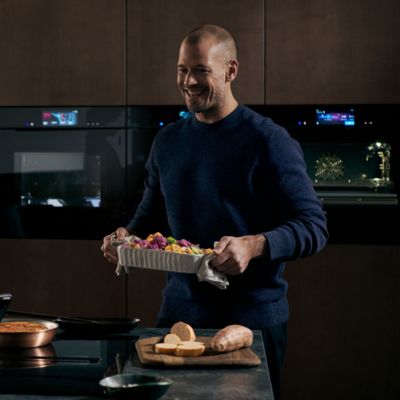 Man holding a baking tray with food, in front of Siemens ovens