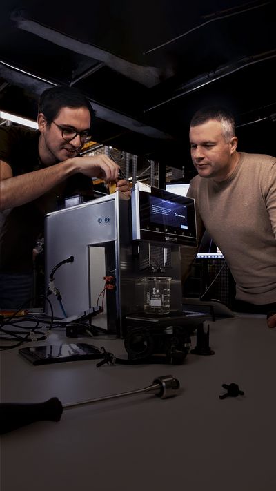 Due uomini in uno stabilimento produttivo lavorano su una macchina automatica per espresso di Siemens - uno di loro sta avvitando la macchina.