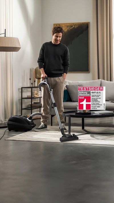 Ein Mann mit schwarzem Pullover und beiger Hose saugt sein Wohnzimmer mit einem Siemens VSZ7X500 Staubsauger mit Beutel. In das Bild ist ein Testsieger Logo der Stiftung Warentest mit dem Testurteil Gut (2,1) für das Gerät integriert.