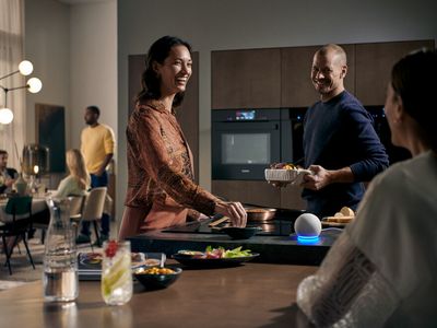 Drei Personen stehen um eine Kochinsel und unterhalten sich. Auf der Kochinsel steht ein Google Sprachassistent. WLAN fähige Siemens Hausgeräte mit Home Connect sind per Sprache steuerbar