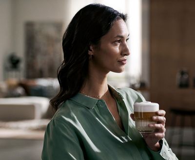 Eine Frau hat ein Glas mit Cappucchino gefüllt in der Hand. Sie sitzt in einer Wohnküche.