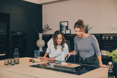 Zwei Frauen in einer modernen Küche bereiten gemeinsam ein Gericht vor. Eine schneidet Gemüse, die andere arrangiert das Essen im Backblech.