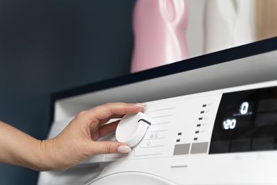 Ręka regulująca pokrętło nowoczesnej pralki, na tle delikatnych różowych i białych elementów dekoracyjnych.