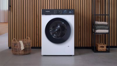 Une machine à laver Siemens moderne avec un écran frontal noir, placée dans une pièce minimaliste avec des accents en bois et des serviettes soigneusement empilées.