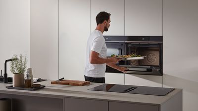 En man i ett ljust modernt kök ställer in en ugnsplåt full med grönsaker i ugnen.