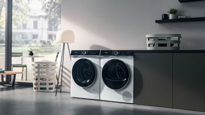 Lavadora y secadora Siemens en una sala de lavandería moderna con grandes ventanales y cesta de ropa blanca.