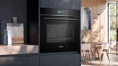 A sleek black Siemens oven with a digital display is integrated into dark kitchen cabinetry, with a modern dining area visible through an open doorway.