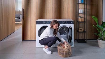 Osoba klęczy obok nowoczesnej pralki i układa ubrania z plecionego kosza na podłodze w stylowej pralni.